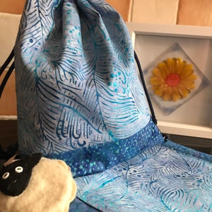 Può includere: Una borsa a cordoncino blu e bianca con un motivo a foglie. La borsa è realizzata in tessuto e ha un bordo blu e bianco. Un piccolo peluche è nella borsa.