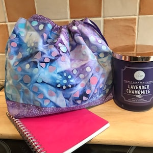 Puede incluir: Una bolsa de cordón de color morado y azul con un cuaderno rosa y una vela perfumada de lavanda y manzanilla. El tarro de la vela es morado con letras doradas y una tapa dorada.