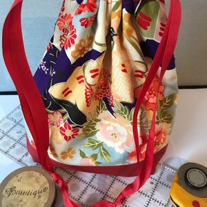 Peut inclure: Un sac à cordon rouge avec un imprimé floral. Le sac a un cordon de serrage en ruban rouge et est fait d'un tissu doux. Le sac est posé sur une surface blanche avec une règle et un cutter rotatif.