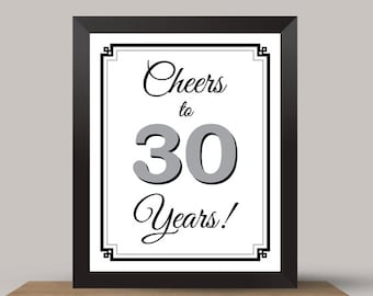 Könnte beinhalten: Ein gerahmter Druck mit dem Text "Cheers to 30 Years!" in schwarzer Schrift und grauen Blockbuchstaben. Der Druck befindet sich in einem schwarzen Rahmen und steht auf einem Holztisch. Der Hintergrund ist hellgrau.