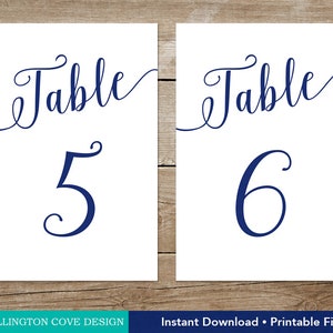 May include: Two white rectangular table number cards. Each card has the word "Table" in a navy blue script font at the top. Below, a large navy blue number, either "5" or "6", is displayed. The cards are designed for weddings or events.