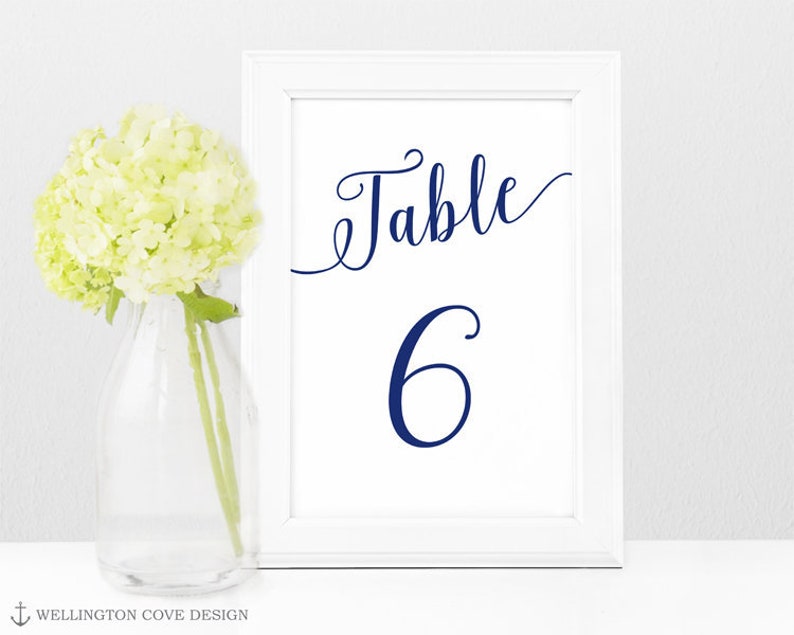 May include: A white framed table number card with the word "Table" and the number "6" in navy blue script. A vase with yellow flowers sits to the left of the card. The card is on a white surface.