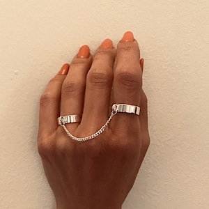 May include: A silver ring set with two bands connected by a chain. The ring is worn on the index and middle fingers.