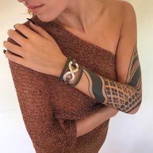 May include: A brown leather bracelet with a silver infinity symbol charm. The bracelet is worn on a person's wrist.