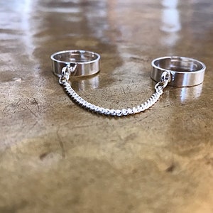 May include: Two silver rings connected by a silver chain. The rings are simple and plain, with no embellishments.