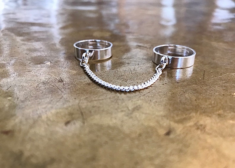 May include: Two silver rings connected by a silver chain. The rings are simple and plain, with no embellishments.