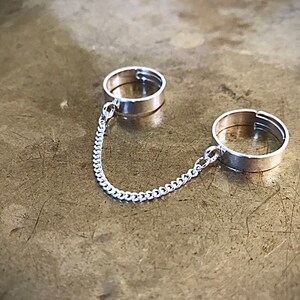 May include: Two silver rings connected by a silver chain. The rings are simple and have a flat band.