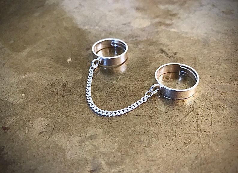 May include: Two silver rings connected by a silver chain. The rings are simple and have a flat band.