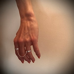 May include: Two silver rings connected by a chain. The rings are worn on the index and middle fingers.