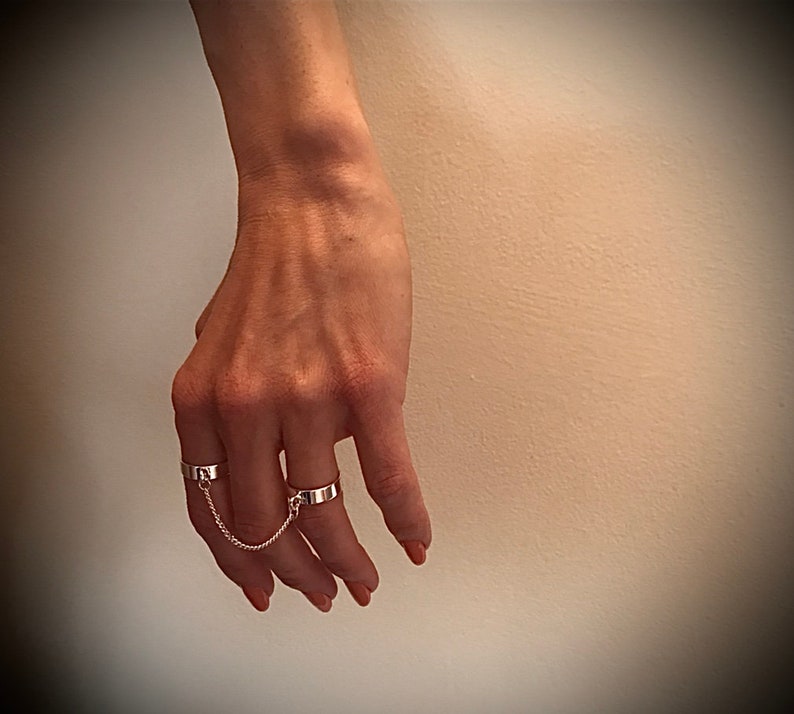 May include: Two silver rings connected by a chain. The rings are worn on the index and middle fingers.
