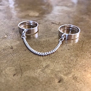 May include: Two silver rings connected by a silver chain. The rings are simple and plain, with no embellishments.