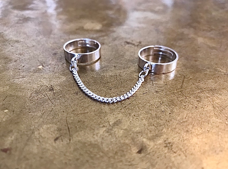 May include: Two silver rings connected by a silver chain. The rings are simple and plain, with no embellishments.