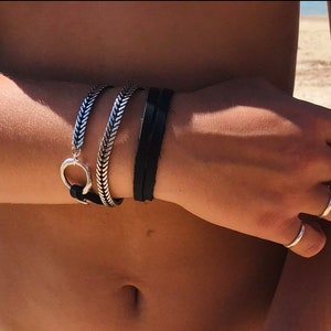 Pode incluir: Uma pessoa usando um bracelete de corrente prateada com um bracelete de couro preto no pulso. Ela também está usando vários anéis de prata nos dedos.