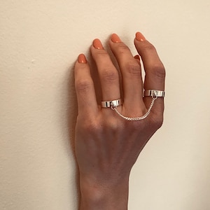 May include: Two silver rings connected by a chain. The rings are worn on the middle and ring fingers of a hand.
