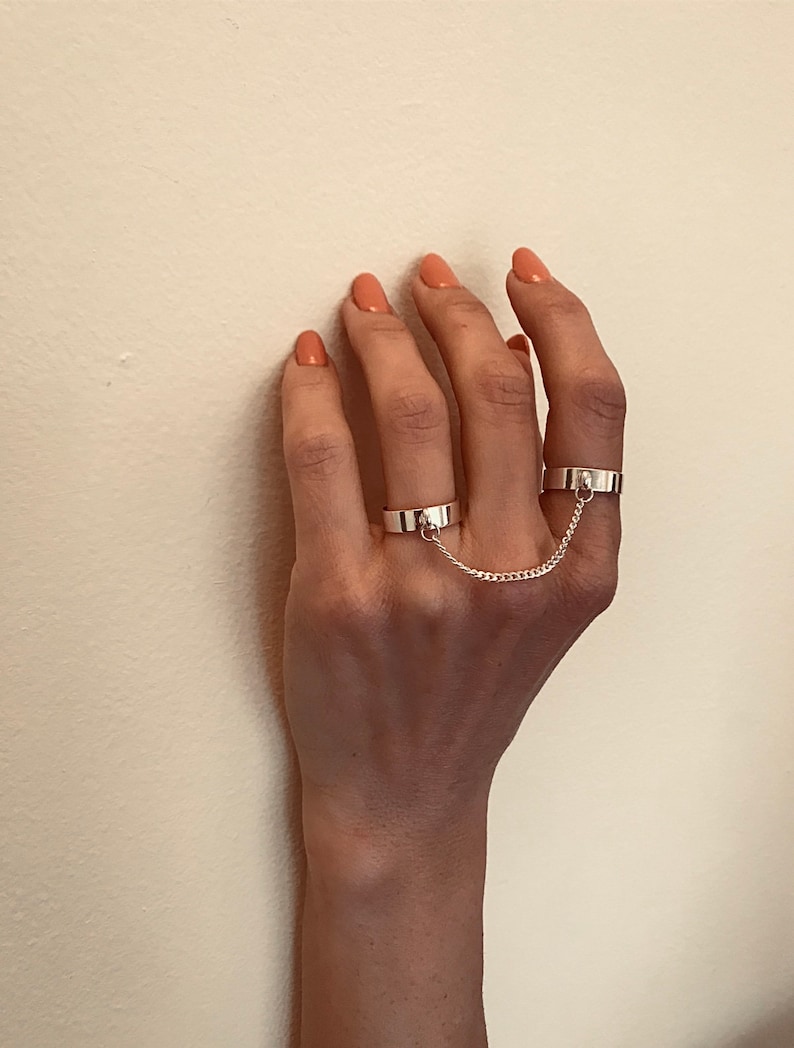 May include: Two silver rings connected by a chain. The rings are worn on the middle and ring fingers of a hand.