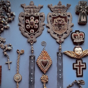 May include: A collection of vintage-style religious ornaments, including a cross, a heart, and a crown. The ornaments are made of gold and silver metal and have a distressed finish. They are hanging from a blue background.