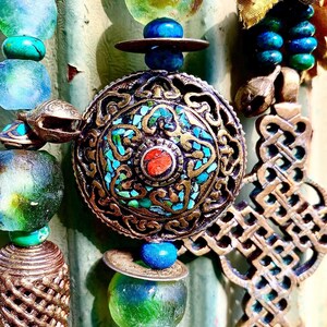 May include: A close-up of a decorative pendant with intricate gold filigree and turquoise and coral accents. The pendant is part of a multi-strand necklace with beads of various colors and textures.