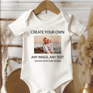 May include: A white baby bodysuit hanging on a wooden hanger. The bodysuit features a printed photo of a man and child. Above the photo, the text reads "CREATE YOUR OWN". Below the photo, the text reads "ANY IMAGE, ANY TEXT" and "UPLOAD YOUR OWN DESIGN."