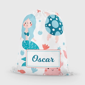 May include: White drawstring bag featuring a mermaid, jellyfish, crab, and fish illustrations in shades of blue, pink, and coral. The name "Oscar" is displayed in a teal font within a white and pink framed label.