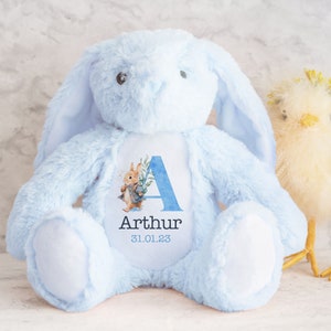 May include: A light blue plush bunny with long ears, a white patch on its chest, and a personalized patch featuring the letter A, the name Arthur, and the date 31.01.23. The bunny is sitting on a white surface next to a yellow chick.