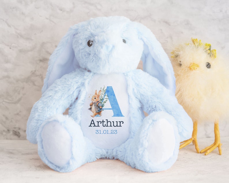 May include: A light blue plush bunny with long ears, a white patch on its chest, and a personalized patch featuring the letter A, the name Arthur, and the date 31.01.23. The bunny is sitting on a white surface next to a yellow chick.