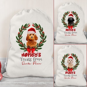 May include: Three white canvas Christmas gift bags with red and green wreaths, pine cones, and a dog wearing a Santa hat. The bags are personalised with the names "Honey's", "Millie's", and "Ruby's" and the text "Treats from Santa Paws".