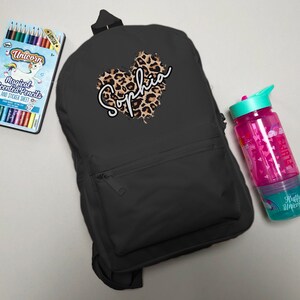 May include: Black backpack with a leopard print heart design and the name "Sophia" in white script. A box of unicorn-themed colored pencils and a pink water bottle with a teal lid are next to the backpack.