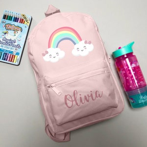 May include: A pink backpack featuring a rainbow and cloud design, personalised with the name "Olivia." Includes a matching water bottle and a box of unicorn-themed coloured pencils. The backpack has a front zip pocket.