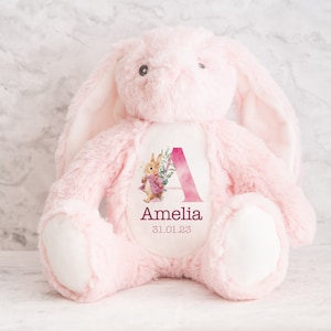 May include: Pink plush bunny with a white patch on its chest. The patch features a watercolor illustration of the letter A with pink flowers and a small bunny. The text "Amelia 31.01.23" is printed below the illustration.