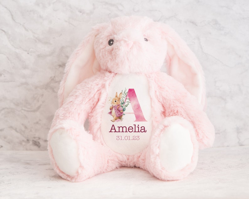 May include: Pink plush bunny with a white patch on its chest. The patch features a watercolor illustration of the letter A with pink flowers and a small bunny. The text "Amelia 31.01.23" is printed below the illustration.