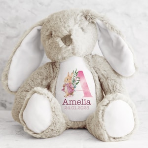 May include: A beige plush bunny with long floppy ears. The bunny has a white patch on its chest with a pink watercolor letter A, floral design, and the name Amelia and date 24.01.2025.