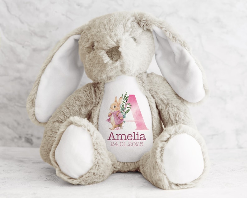 May include: A beige plush bunny with long floppy ears. The bunny has a white patch on its chest with a pink watercolor letter A, floral design, and the name Amelia and date 24.01.2025.