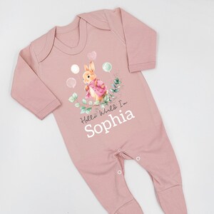 K&ouml;nnte beinhalten: Rosaer Baby Strampler mit einer Peter Rabbit Illustration, Ballons und dem Text "Hello World I'm Sophia".