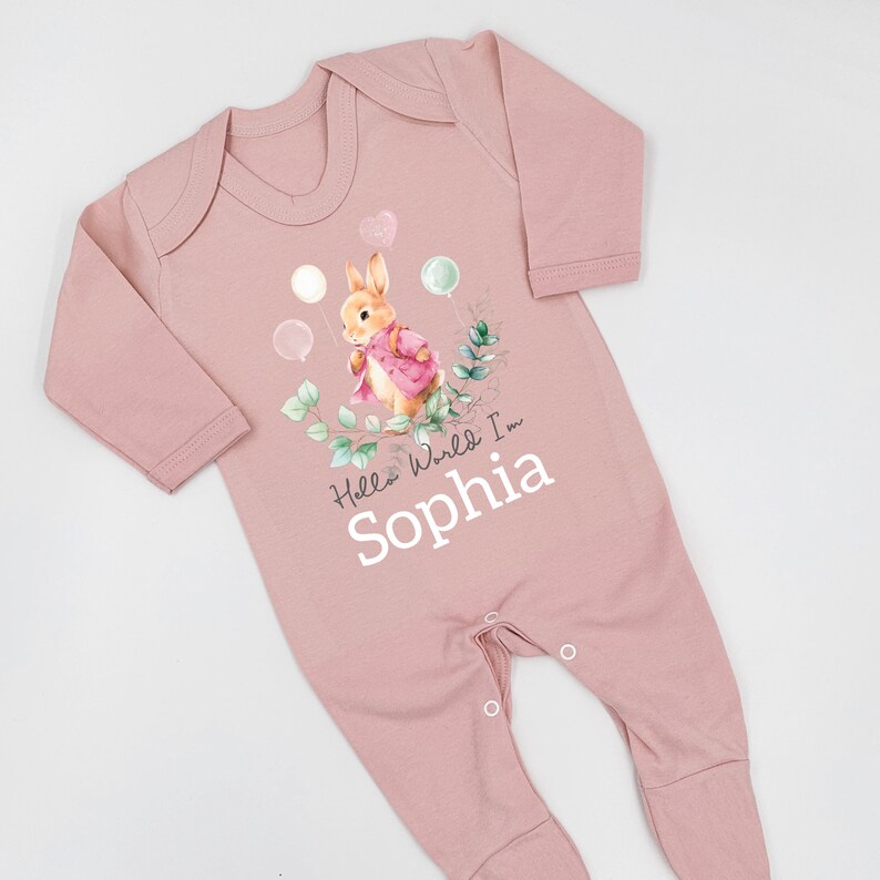 K&ouml;nnte beinhalten: Rosaer Baby Strampler mit einer Peter Rabbit Illustration, Ballons und dem Text "Hello World I'm Sophia".