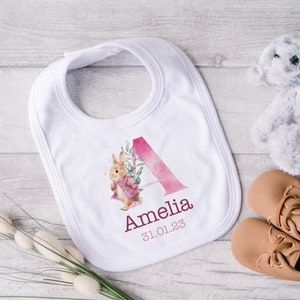 May include: White baby bib with a pink watercolor letter A, a floral design, and a rabbit illustration. The bib is personalized with the name Amelia and the date 31.01.23.