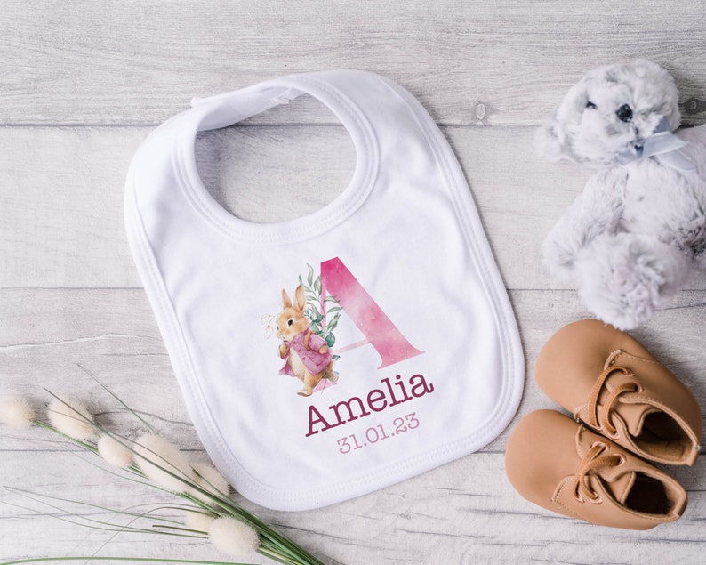 May include: White baby bib with a pink watercolor letter A, a floral design, and a rabbit illustration. The bib is personalized with the name Amelia and the date 31.01.23.