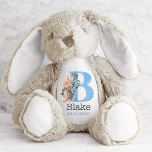 May include: A beige plush bunny with long ears and a white patch on its chest. The patch features a blue letter "B" with a green leaf and a watercolor illustration of Peter Rabbit. The text "Blake 24.01.2025" is printed below the letter.