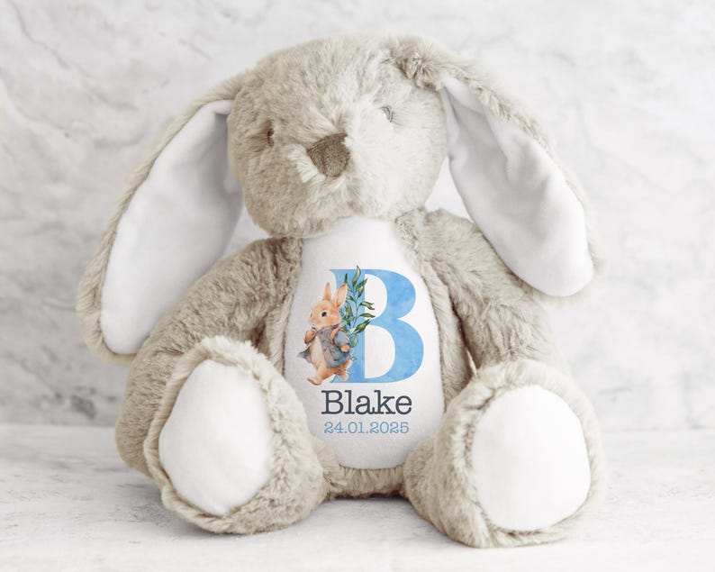 May include: A beige plush bunny with long ears and a white patch on its chest. The patch features a blue letter "B" with a green leaf and a watercolor illustration of Peter Rabbit. The text "Blake 24.01.2025" is printed below the letter.