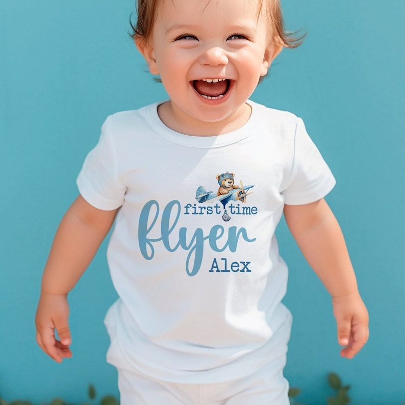 Pu&ograve; includere: Un body bianco per neonato con un aereo blu e un orsacchiotto con un cappello da pilota. Il testo sul body recita "first time flyer Alex".