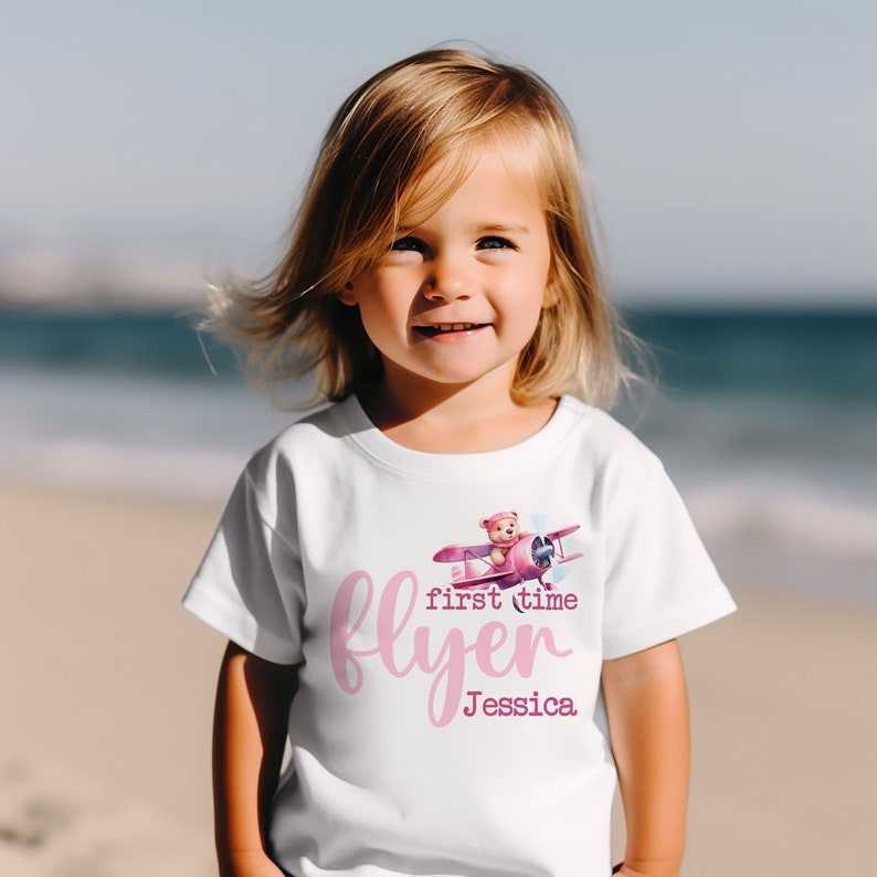 May include: White t-shirt with a pink graphic of a teddy bear flying a pink aeroplane. The text "first time flyer Jessica" is printed in pink on the shirt.
