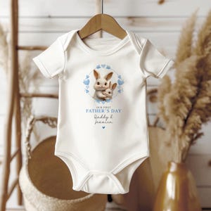 Peut inclure: Combinaison blanche pour b&eacute;b&eacute; avec une bordure de c&oelig;urs bleus autour de deux lapins qui se font un c&acirc;lin. Le texte dit "Notre premi&egrave;re f&ecirc;te des p&egrave;res Papa & Jessica".