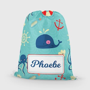 May include: A turquoise blue drawstring bag with a nautical theme. The bag features a whale, octopus, anchor, and a wheel. The bag has a white label with the name "Phoebe" written in black.