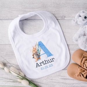 May include: A white baby bib with a blue watercolor letter A, a rabbit illustration, and the name Arthur and date 31.01.23 printed on it.