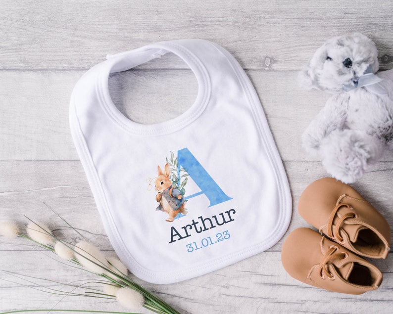 May include: A white baby bib with a blue watercolor letter A, a rabbit illustration, and the name Arthur and date 31.01.23 printed on it.