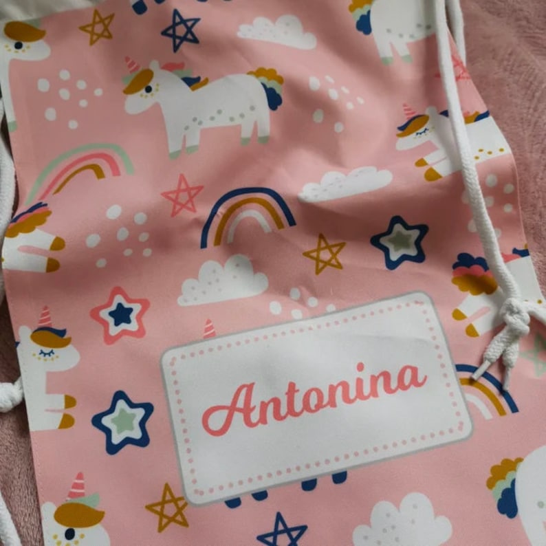 May include: Pink drawstring bag with a white unicorn and rainbow pattern. The bag has a white label with the name Antonina.