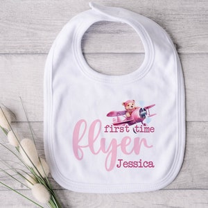 May include: White baby bib with a pink airplane and teddy bear design. The text "first time flyer Jessica" is printed on the bib.