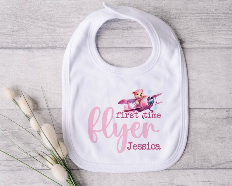 May include: White baby bib with a pink airplane and teddy bear design. The text "first time flyer Jessica" is printed on the bib.