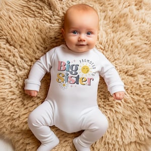 May include: A white baby onesie with a colorful graphic that says "Promoted to Big Sister" with a sun and flowers.