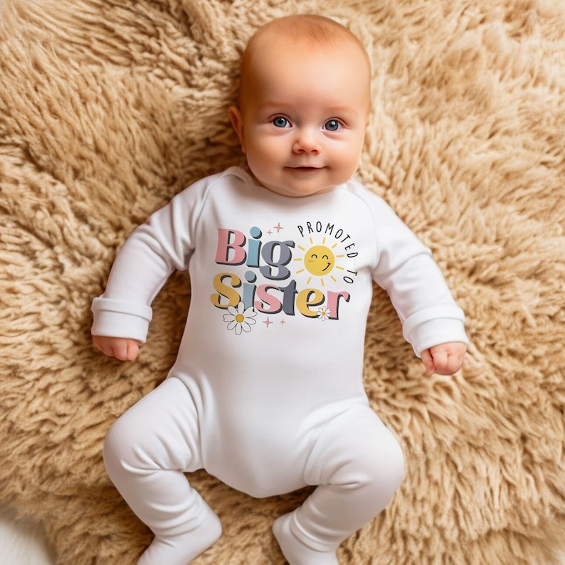May include: A white baby onesie with a colorful graphic that says "Promoted to Big Sister" with a sun and flowers.