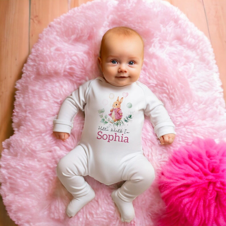 K&ouml;nnte beinhalten: Ein wei&szlig;er Baby-Strampler mit langen &Auml;rmeln zeigt eine Aquarell-Illustration eines Kaninchens, das Ballons h&auml;lt. Der Strampler hat den Text "Hello World I'm Sophia" auf der Vorderseite.